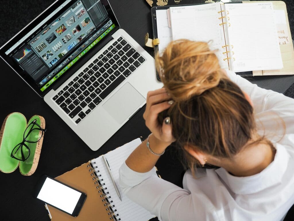 Overhead view of a stressed woman working at a desk with a laptop, phone, and notebooks.